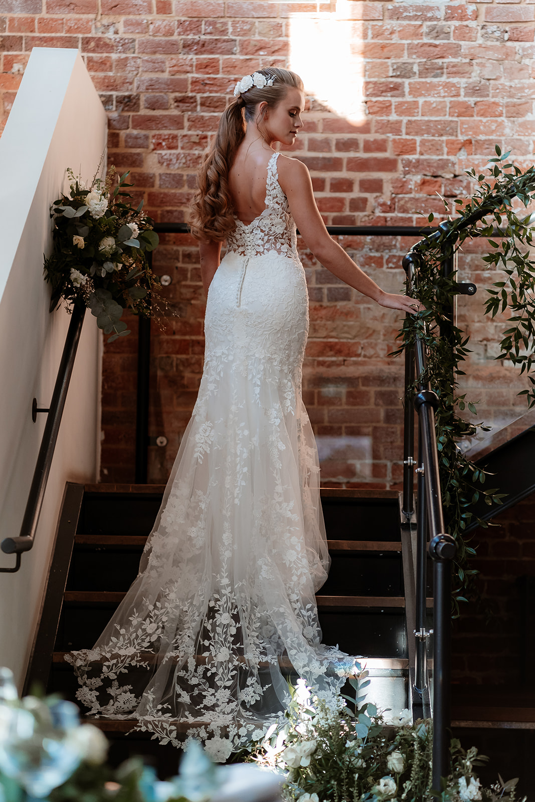 Bride in a white lace fit and flare dress stands on a staircase with her back to the camera. 