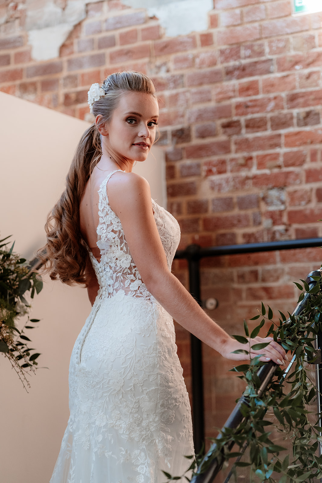 Bride in a white lace dress and bridal ponytail with white flowers in her hair stands on a foliage draped staircase. 