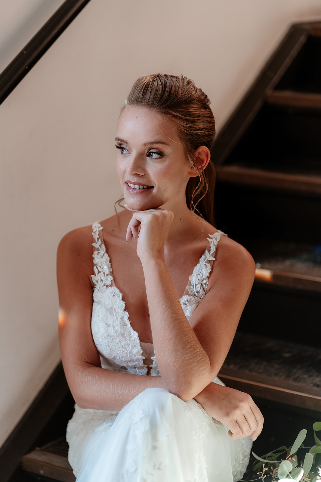 Bride in a white lace dress and bridal ponytail with white flowers in her hair sits on a foliage draped staircase. 