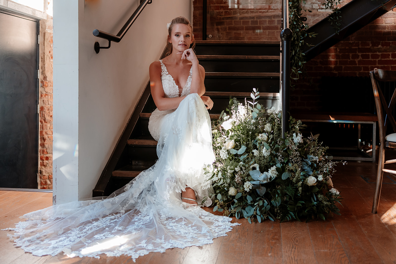 Bride in a white lace dress and bridal ponytail with white flowers in her hair sits on a foliage draped staircase. 