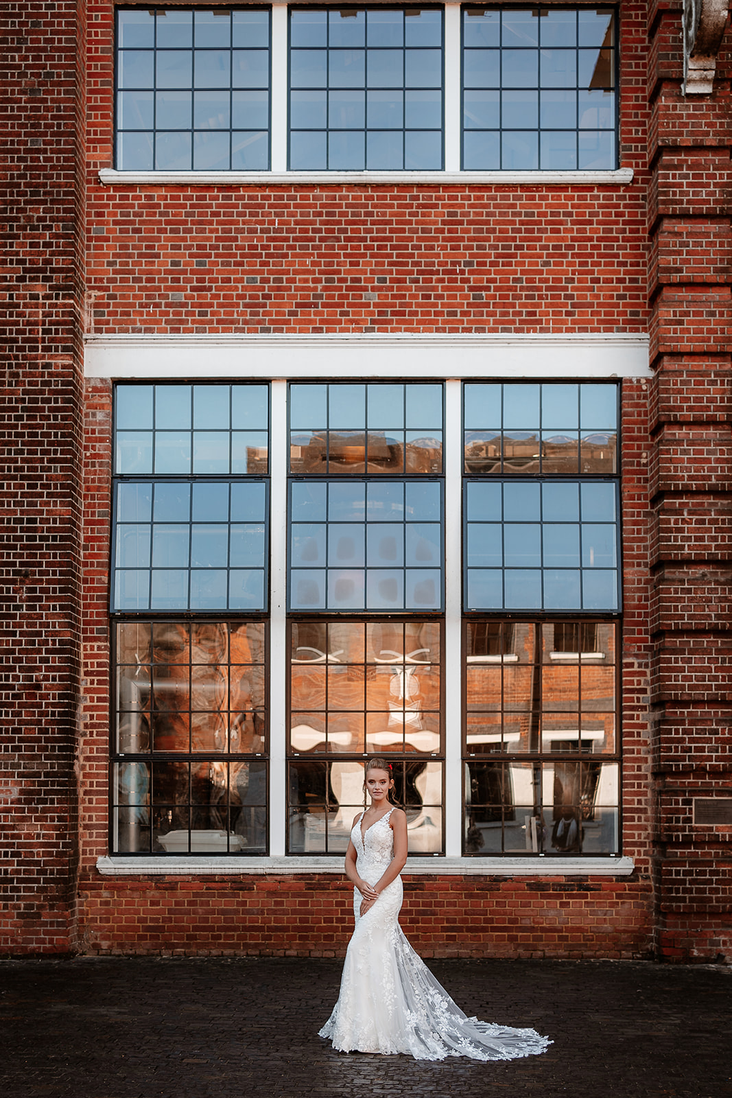 Bridal model in a white lace dress stands in front of tall industrial windows at the Bombay Sapphire Distillery. 