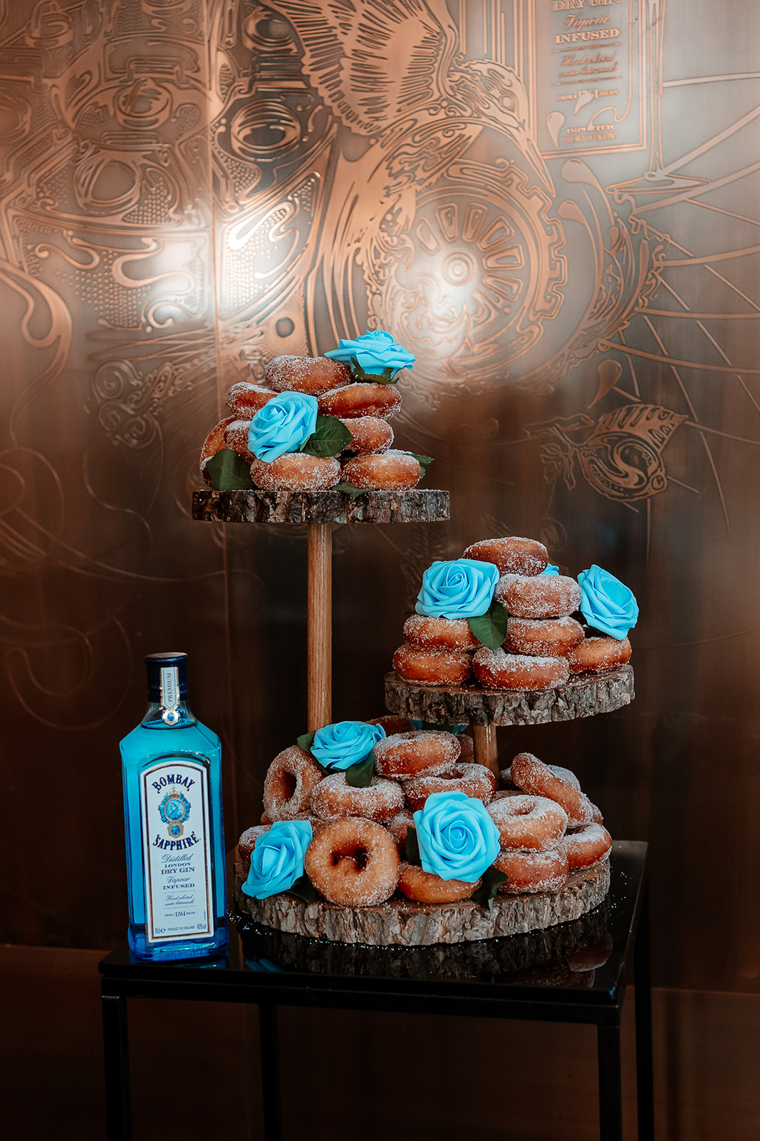 A tower of sugar covered ring doughnuts decorated with blue icing sugar roses sits on a three tier wooden cake stand.