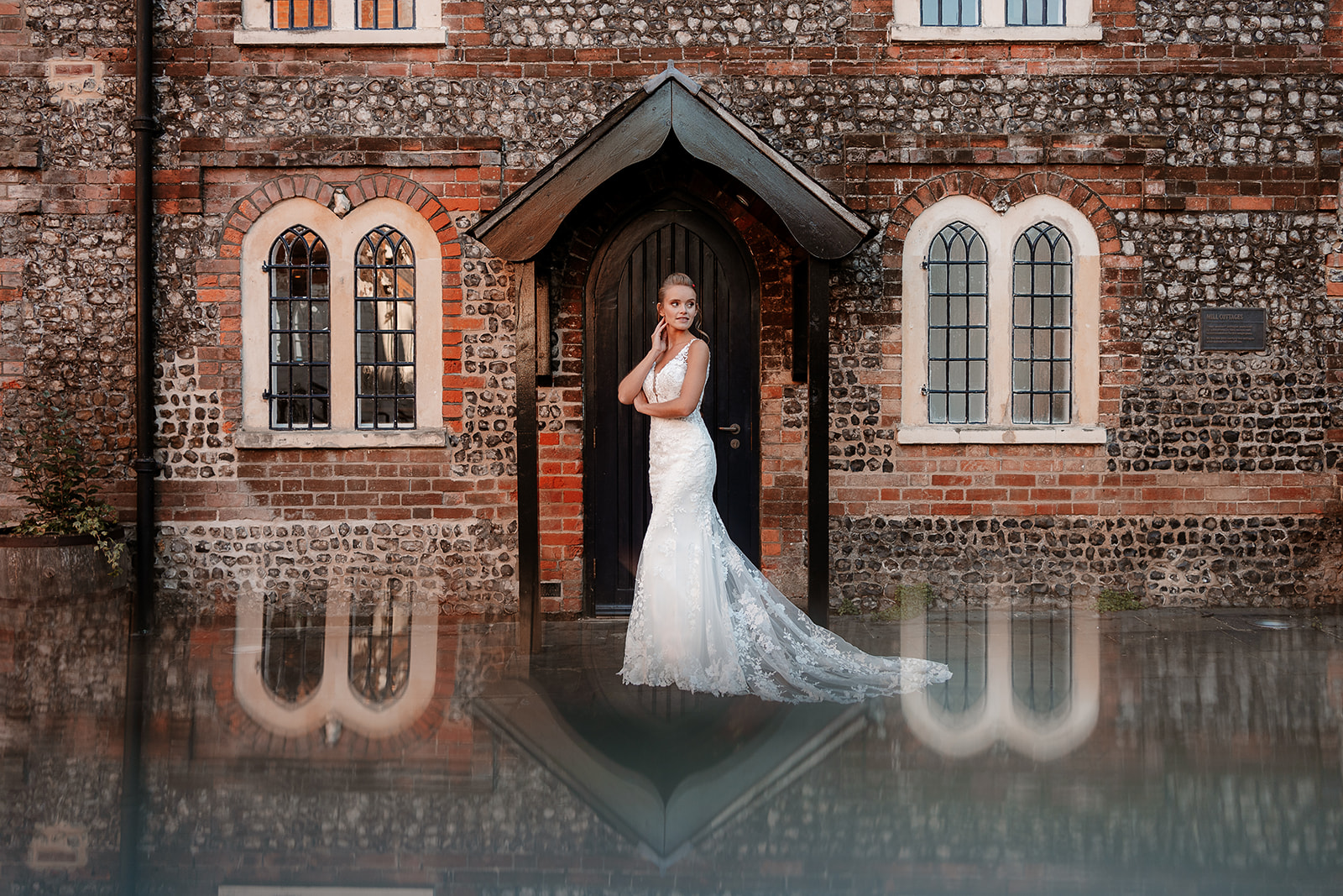 Bride in a white lace dress stands under an old porch, the arched windows and flint walls reflected below her. 