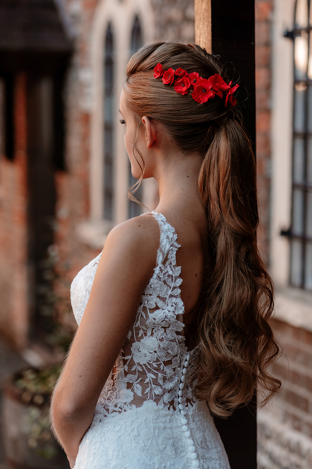 Back view of a bride in a lace wedding dress with red flowers in her ponytail. 