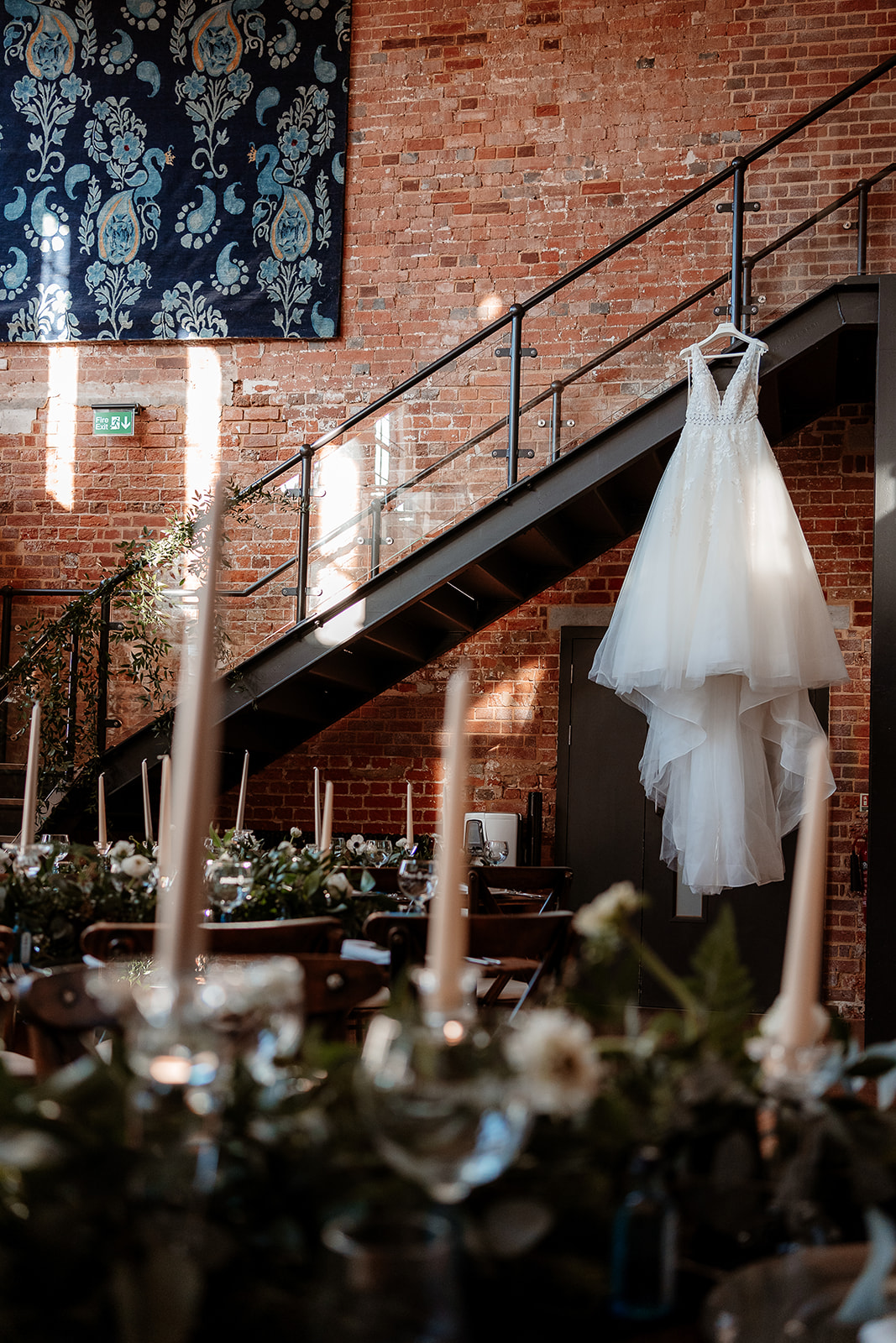 A white bridal ballgown hangs from a staircase with tables laid for a wedding breakfast in the foreground. 