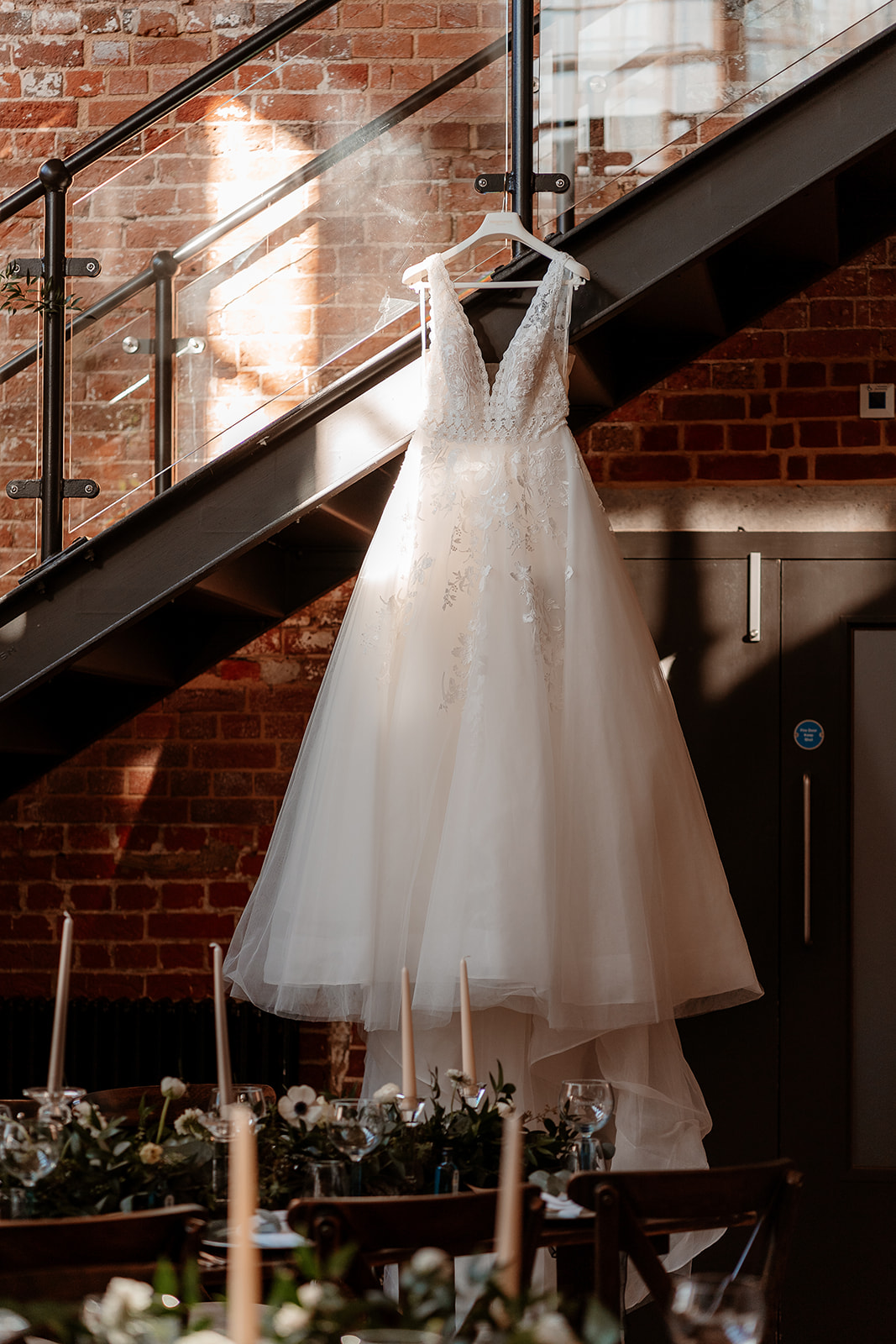 A white bridal ballgown hangs from a staircase with tables laid for a wedding breakfast in the foreground. 