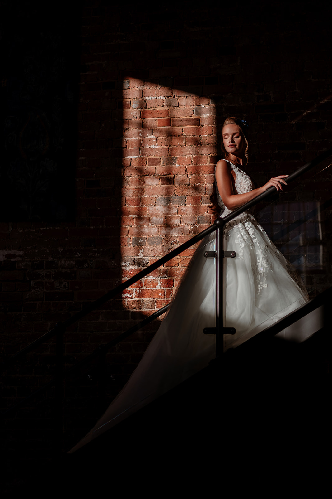 A bride in a wedding dress stands on the stairs in a pocket of bright window light at the Bombay Sapphire Distillery. 