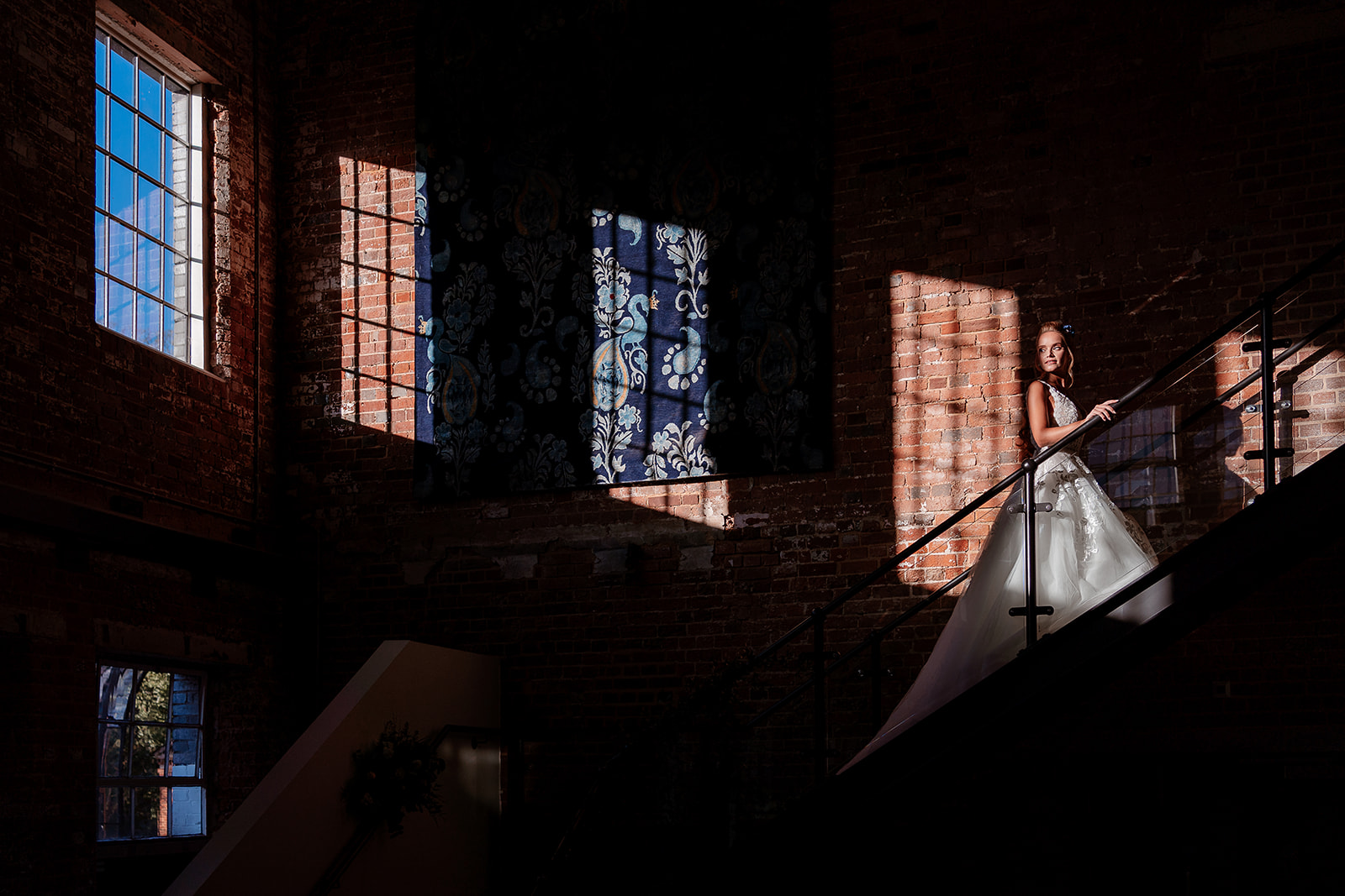 A bride in a wedding dress stands on the stairs in a pocket of bright window light at the Bombay Sapphire Distillery. 