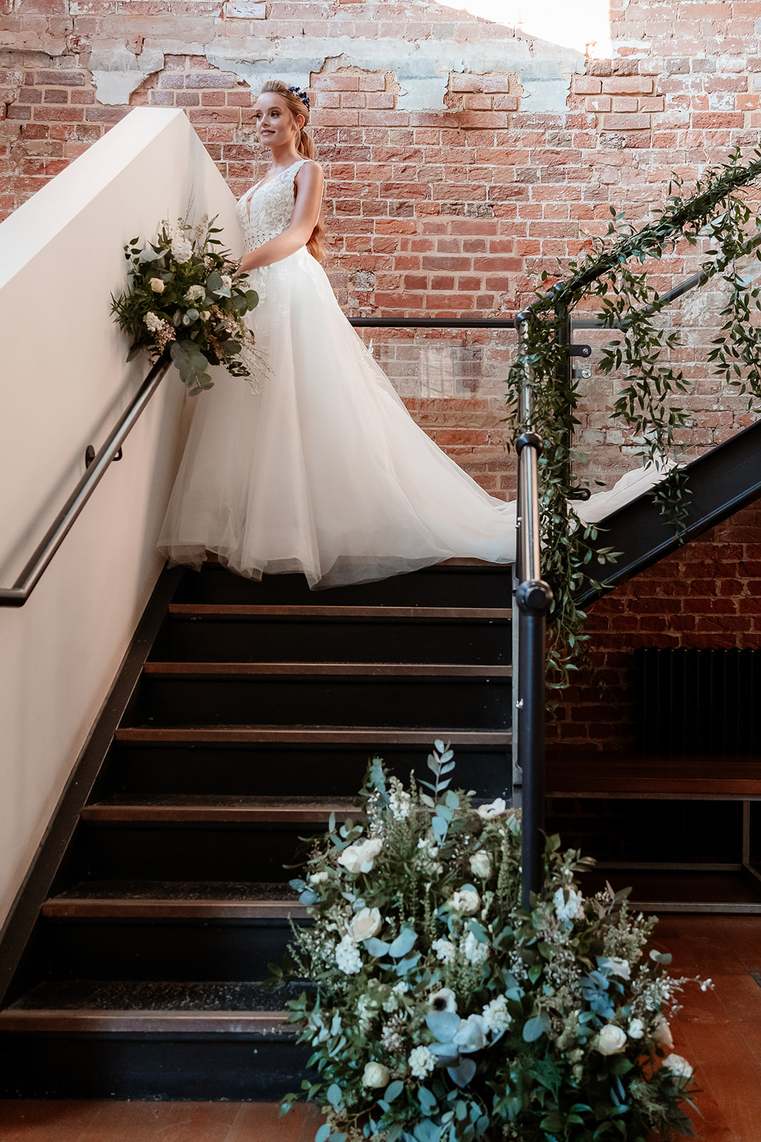 A bride in a white lace wedding dress with her hair in a ponytail stands at a foliage covered staircase. 