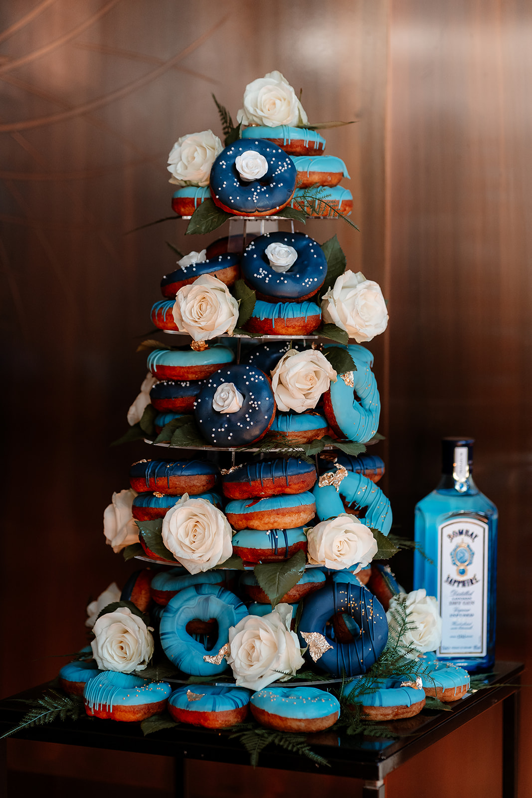 A tower of ring doughnuts decorated with blue icing and white roses next to a bottle of Bombay Sapphire gin. 