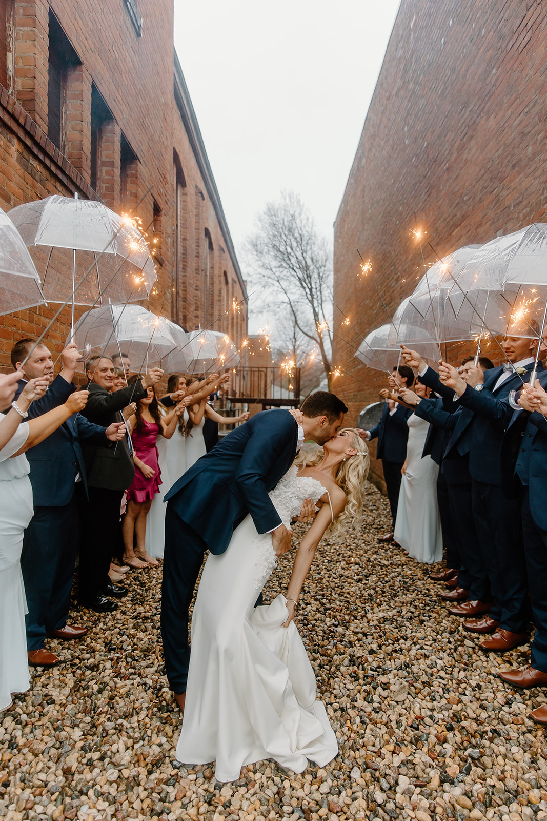 Bride and groom kiss while all of their family and bridal party hold sparklers and umbrellas. 