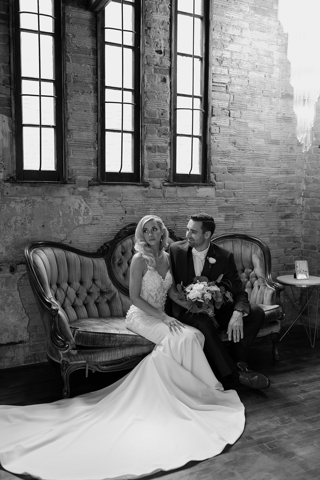 Bride looks off into distance while groom looks at her sitting on a vintage couch. 