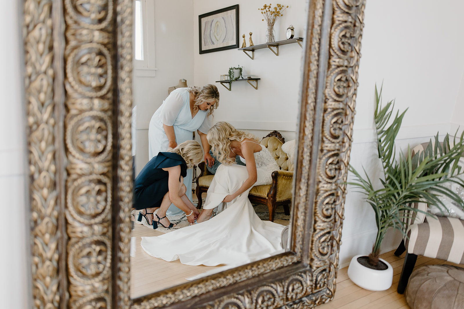 Bride's mom and sister help her put her shoes on as we look in on them through the mirror.