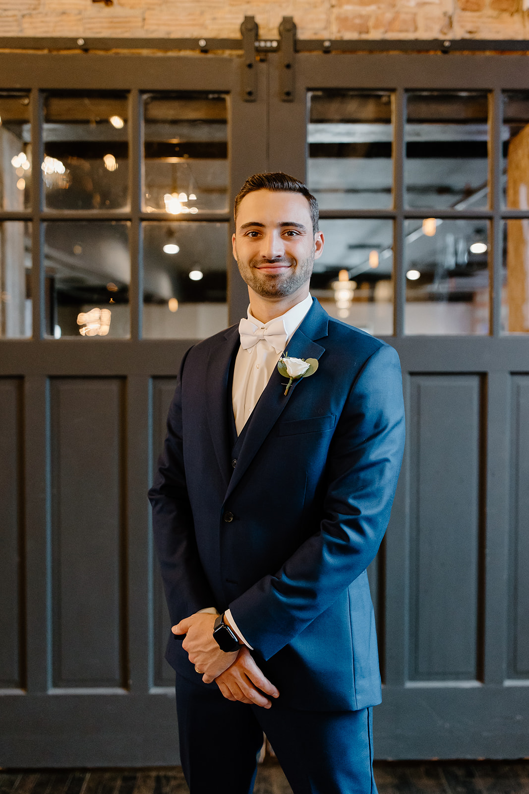 Groom smiles in front of a rolling door and twinkly lights. 