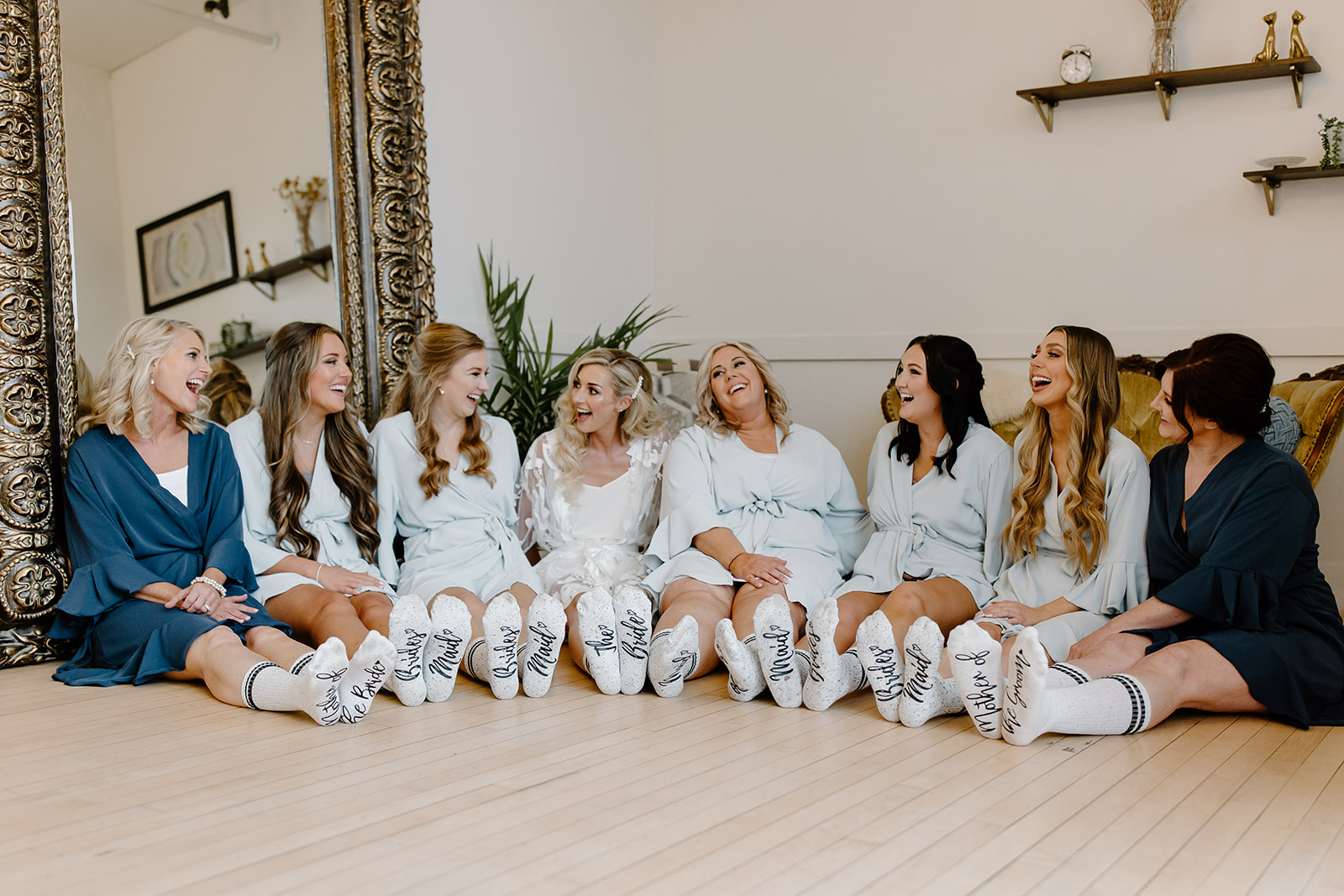 The bride, her bridesmaids, her mom, and her mother in law sit on the floor in their getting ready outfits and socks.