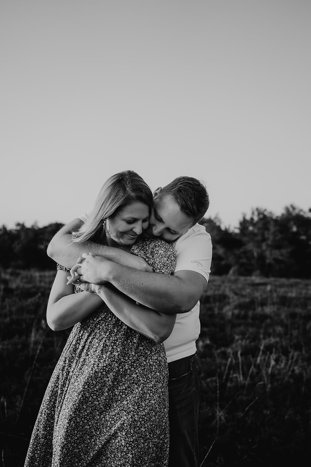 Black and white photo of a man stood behind his fiancée with his arms wrapped around her in a hug