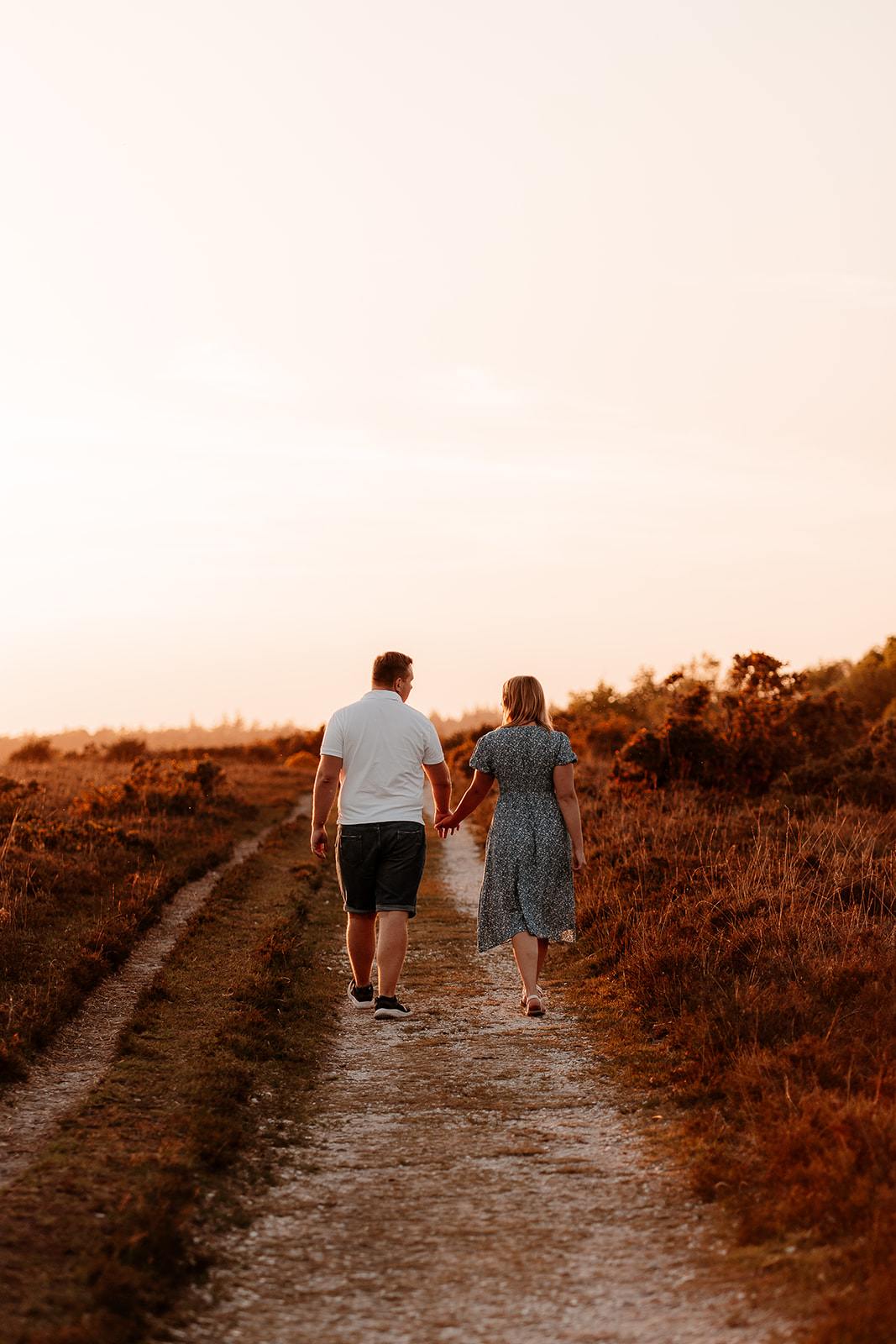 A newly engaged couple walking away from the camera hand in hand towards the sunset. 