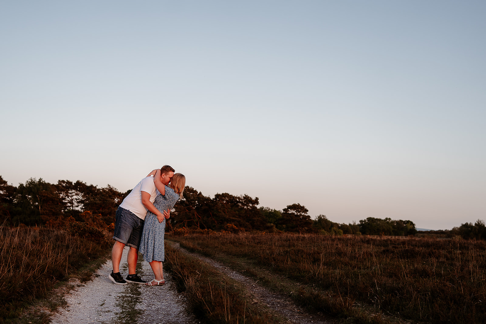 A man dips his new fiancée in a passionate kiss at their engagement photoshoot in the New Forest at sunset. 