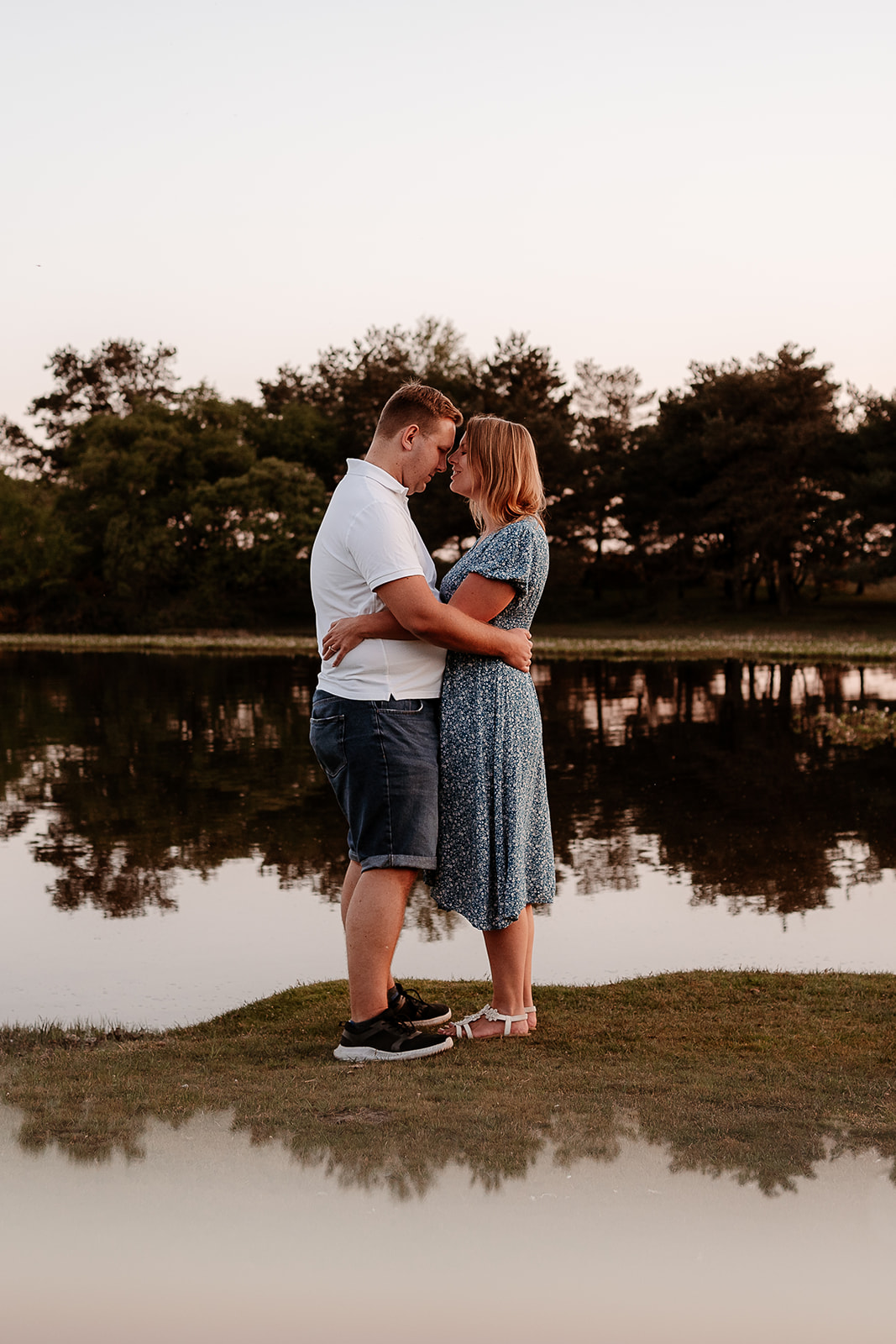 Engagement photoshoot in the New Forest - a couple stand cuddling in front of a scenic pond. 