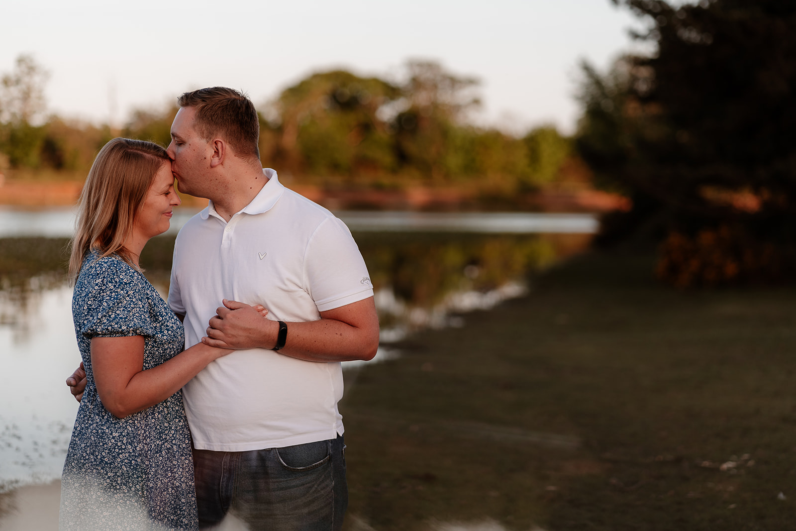 A newly engaged couple embrace in front of a pond in the New Forest on their engagement photoshoot. 