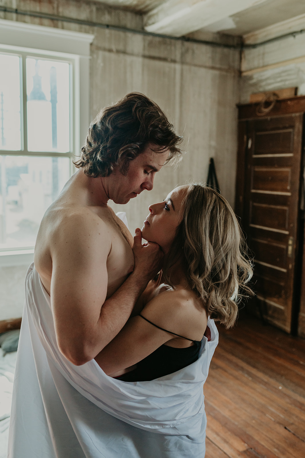 A couple wraps up in a white sheet in an indoor loft space engagement session in Vermilion Alberta.