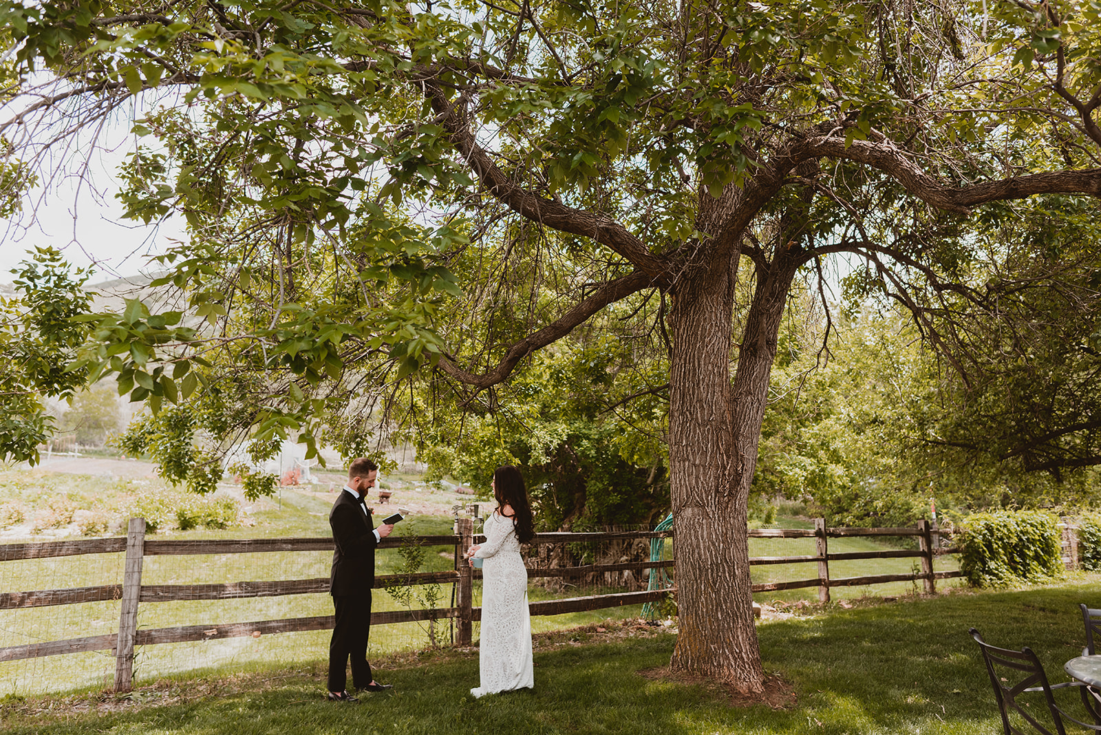 Boho bride and groom stand beneath a large tree next to farm land as they exchange vows in Denver Colorado at Lyons Farm