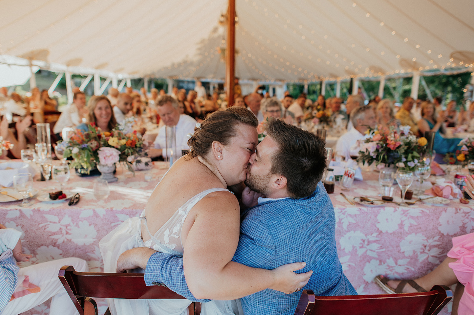 Bride and groom kiss during reception with guests behind them in the tent