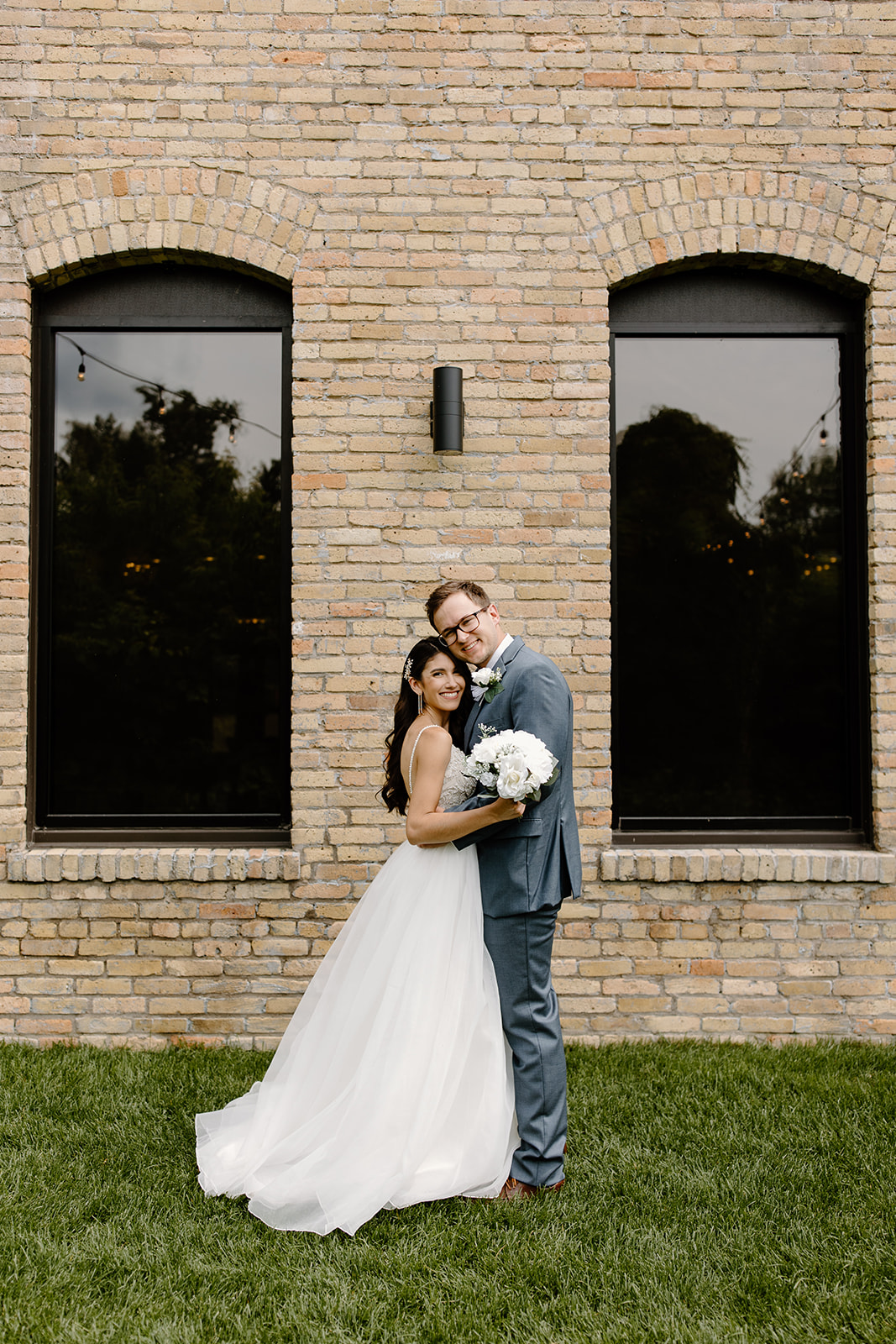 Bride and groom smile in front of a brick wall with black windows
