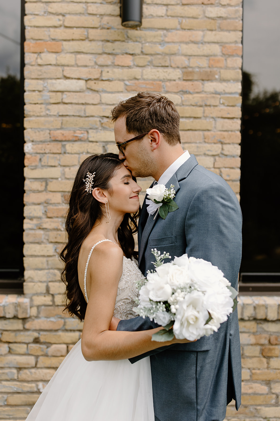 Groom kisses his bride in front of a brick wall with black windows