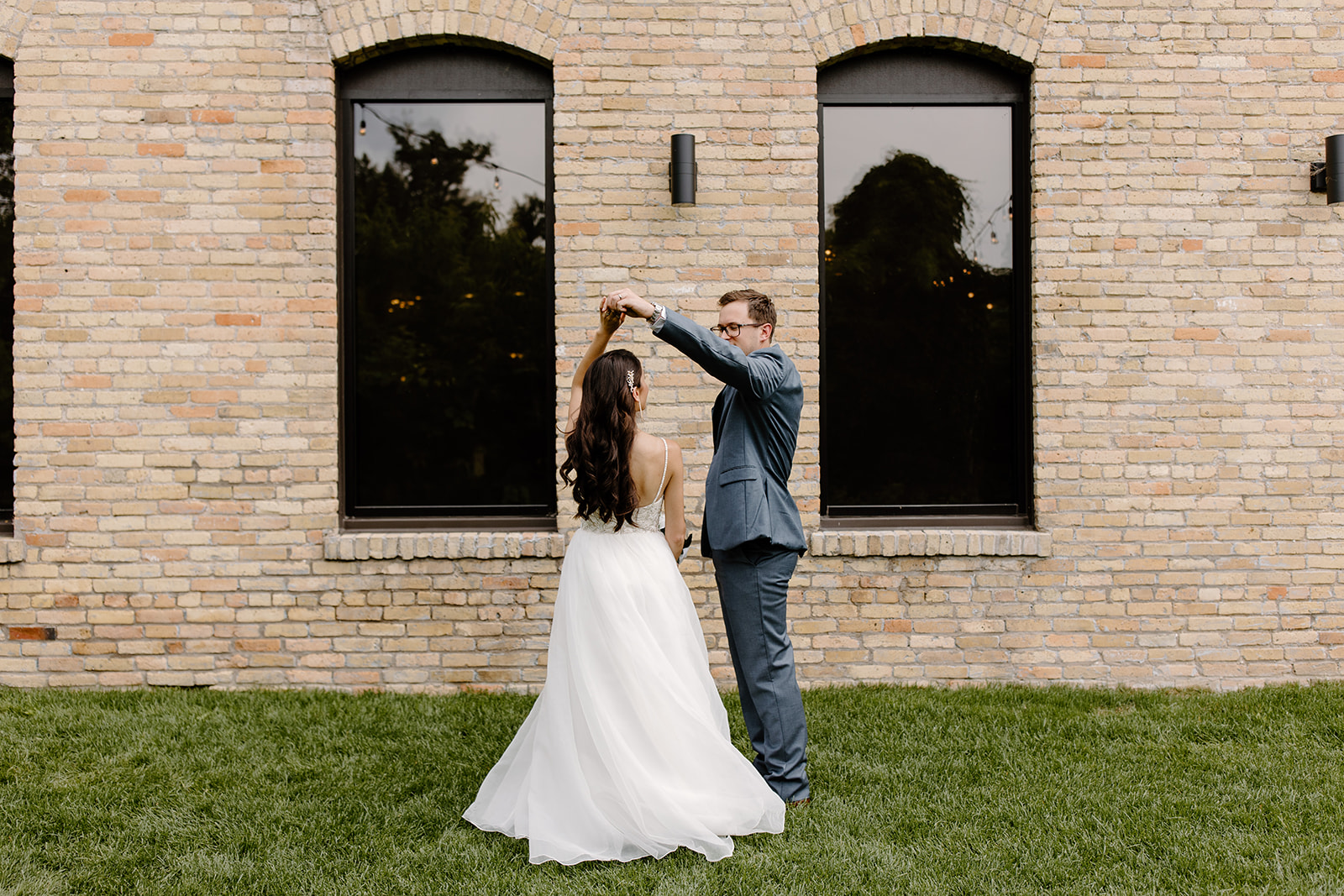 Groom twirls his bride in front of a brick wall with black windows