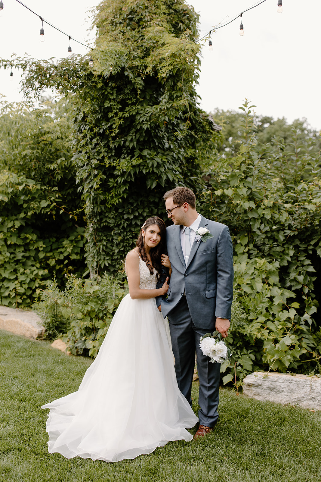 Groom looks down at his bride as she hugs his arm in front of a wall of vines