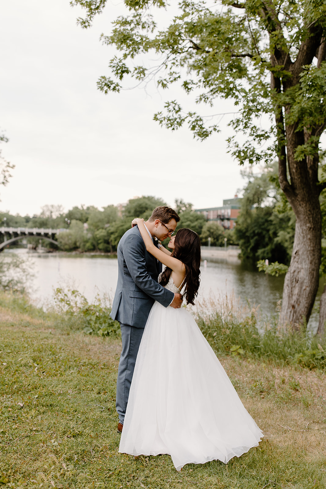Bride has her arms around her groom's neck in front of a river