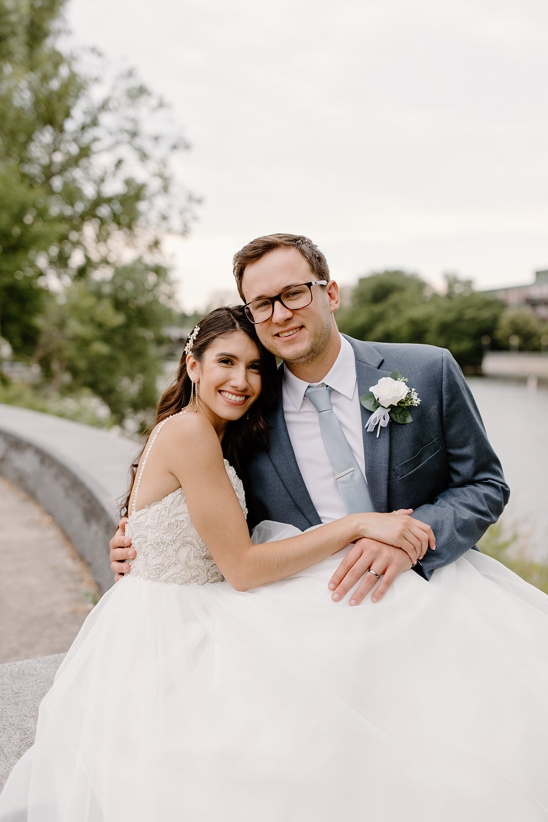 Bride and groom smile while sitting in front of a river