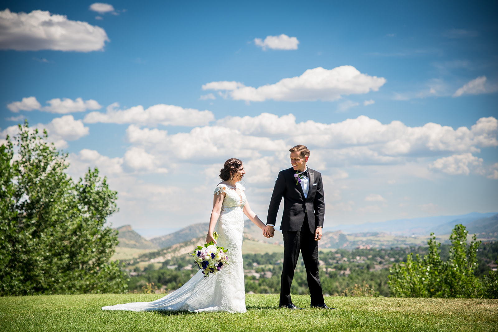 A groom leads his bride across a field of grass with mountain views in the background.