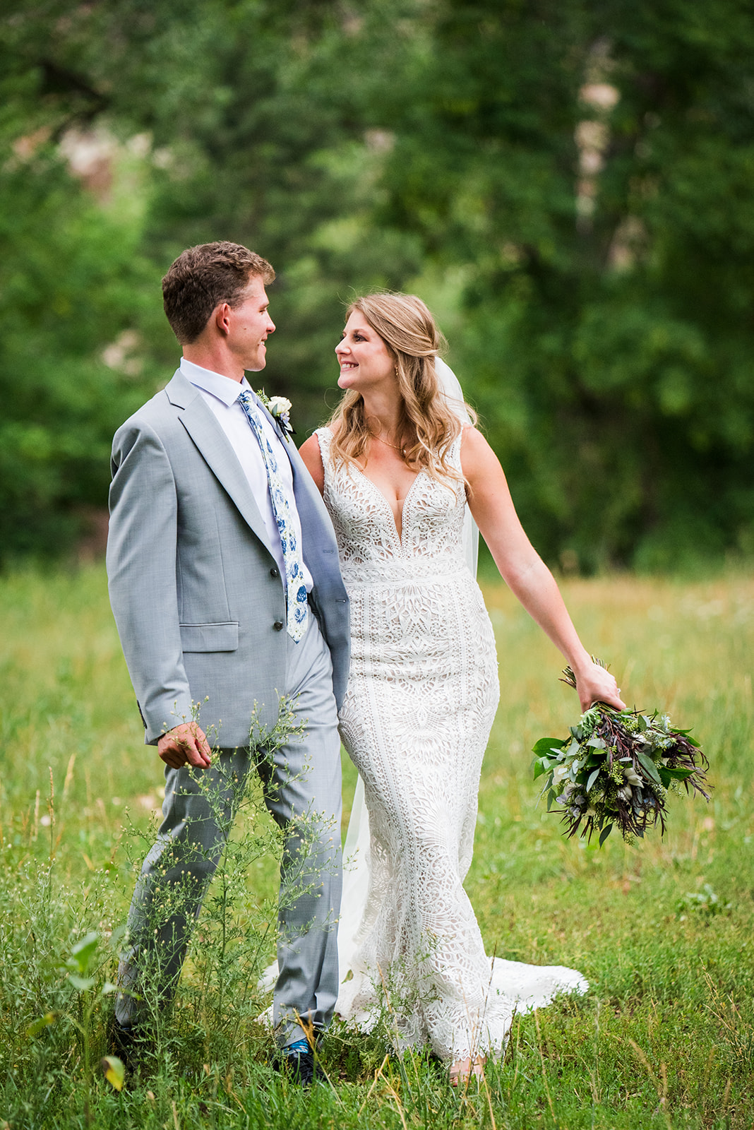 Bride and groom walk through a grassy field hand-in-hand and smile at eachother.