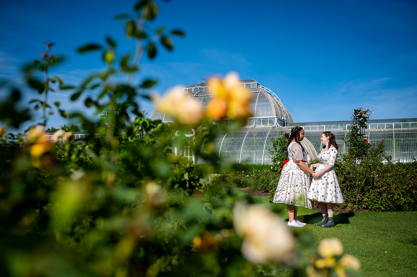Two brides standing in front of the Palm House at Kew Gardens on their wedding day.