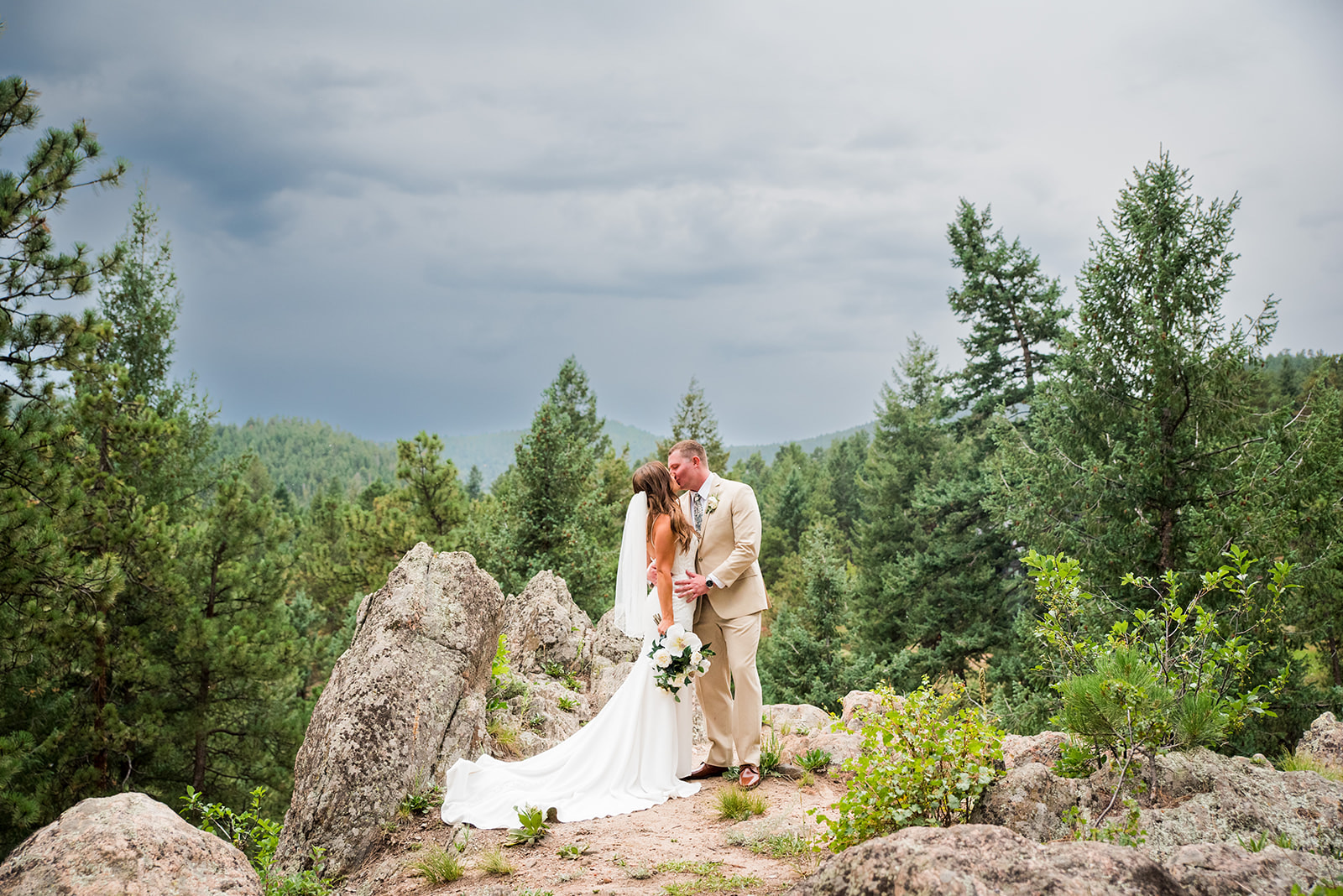 Bride and groom share a kiss on a mountain edge with greenery in the background.