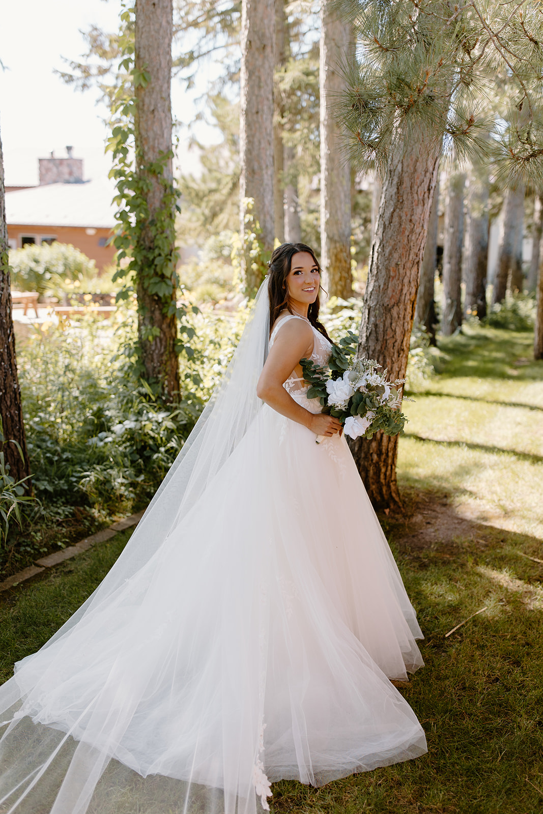 Bride smiles at the camera while holding her bouquet in front of a line of trees