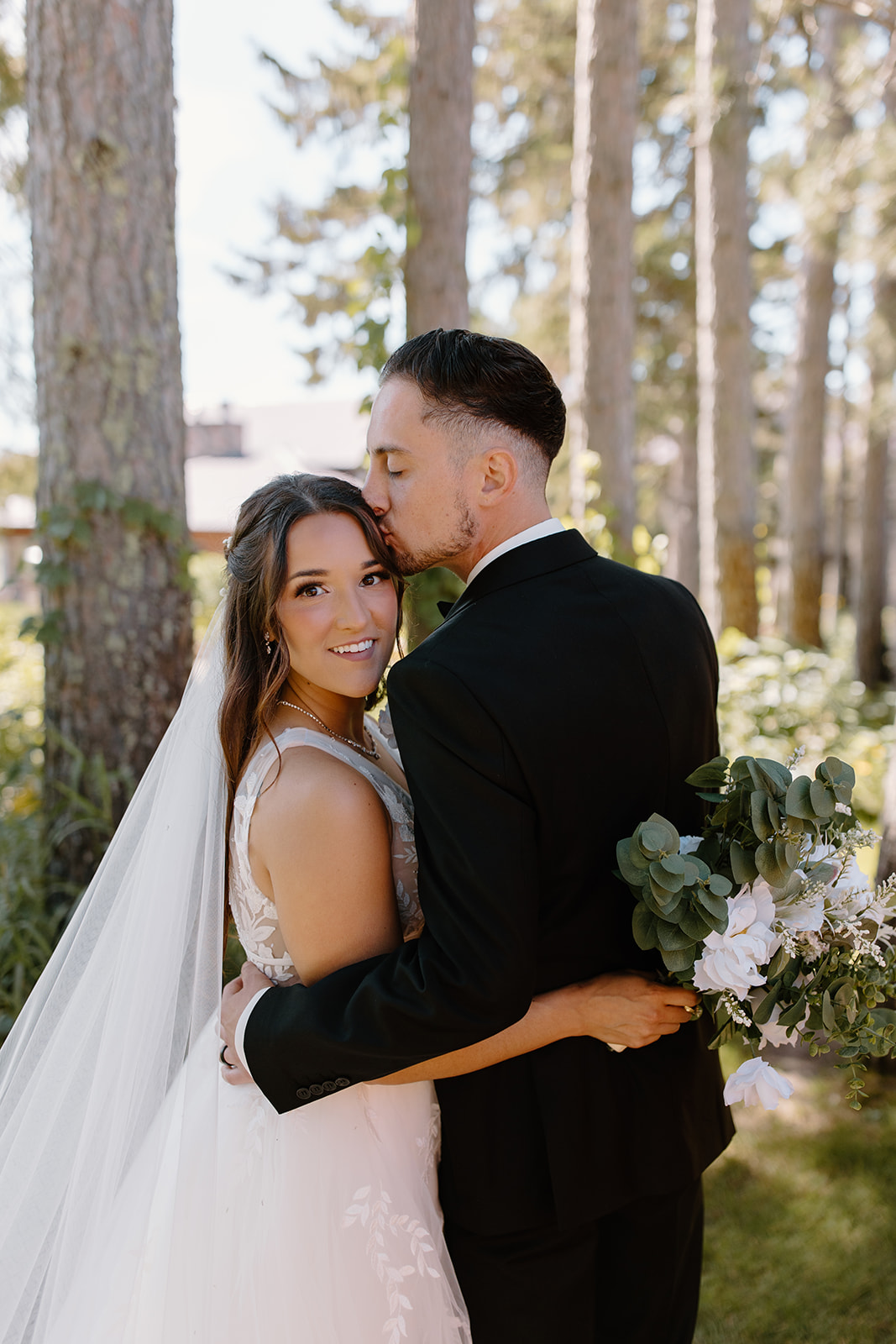 Groom kisses his bride in front of a line of trees