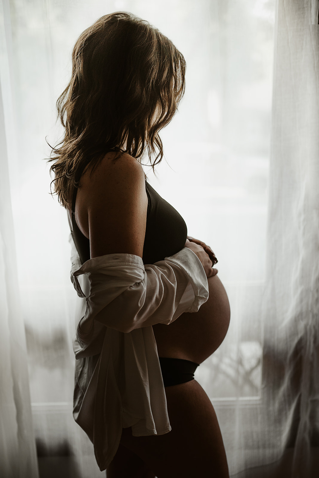 New mother with her pregnant belly standing in a white shirt by window light