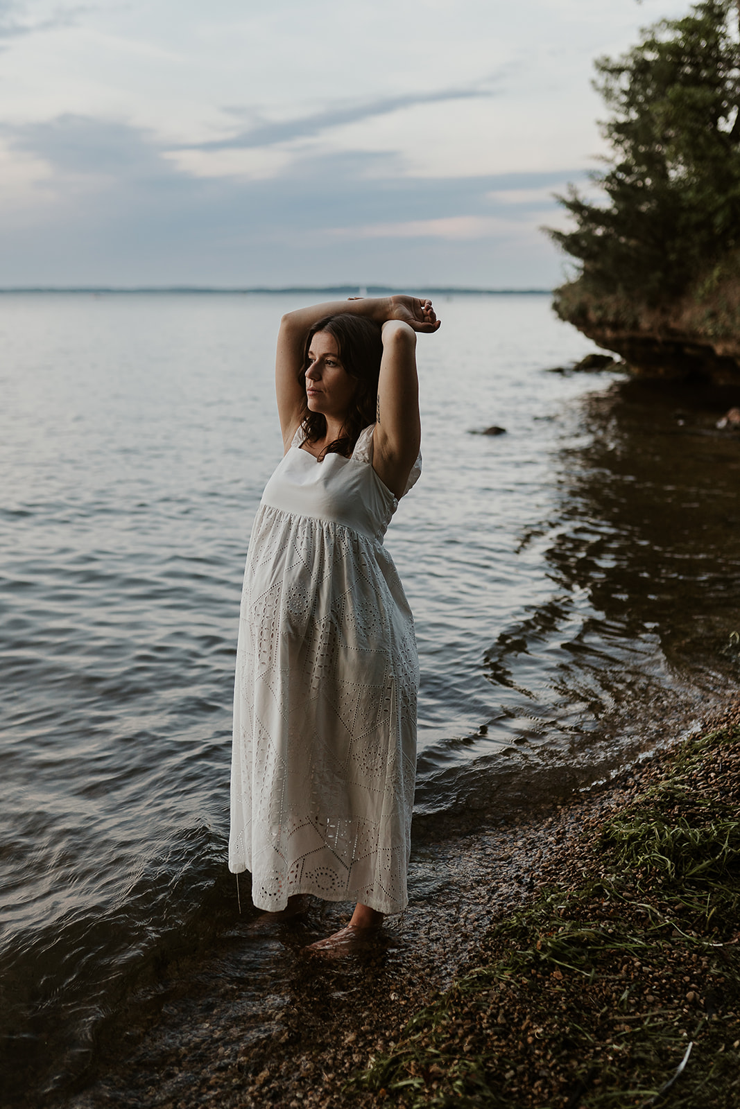 Pregnant mother in a white dress standing in the lakeshore water after sunset