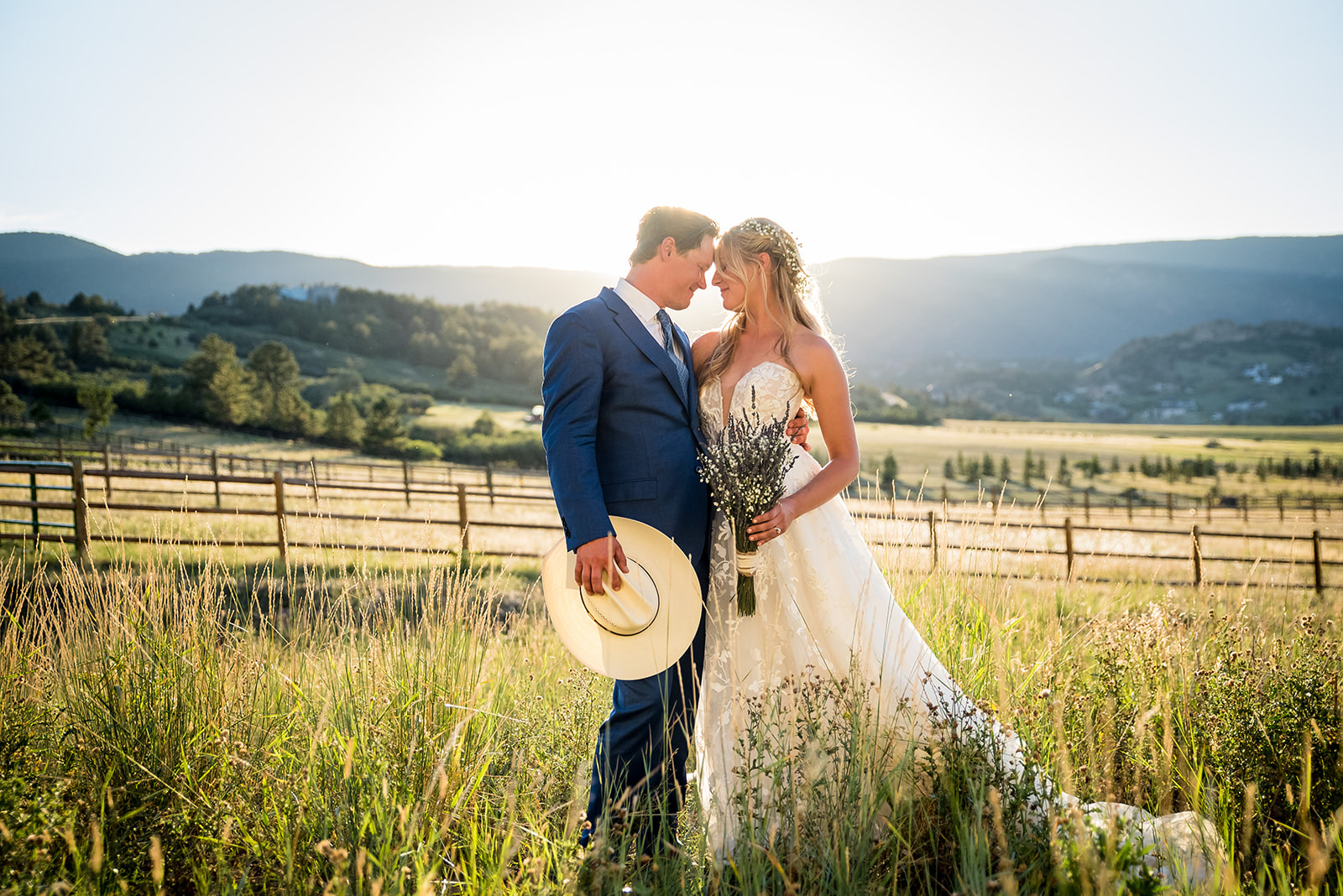 Bride and groom stand in a green pasture with mountain views and the sun setting behind them.
