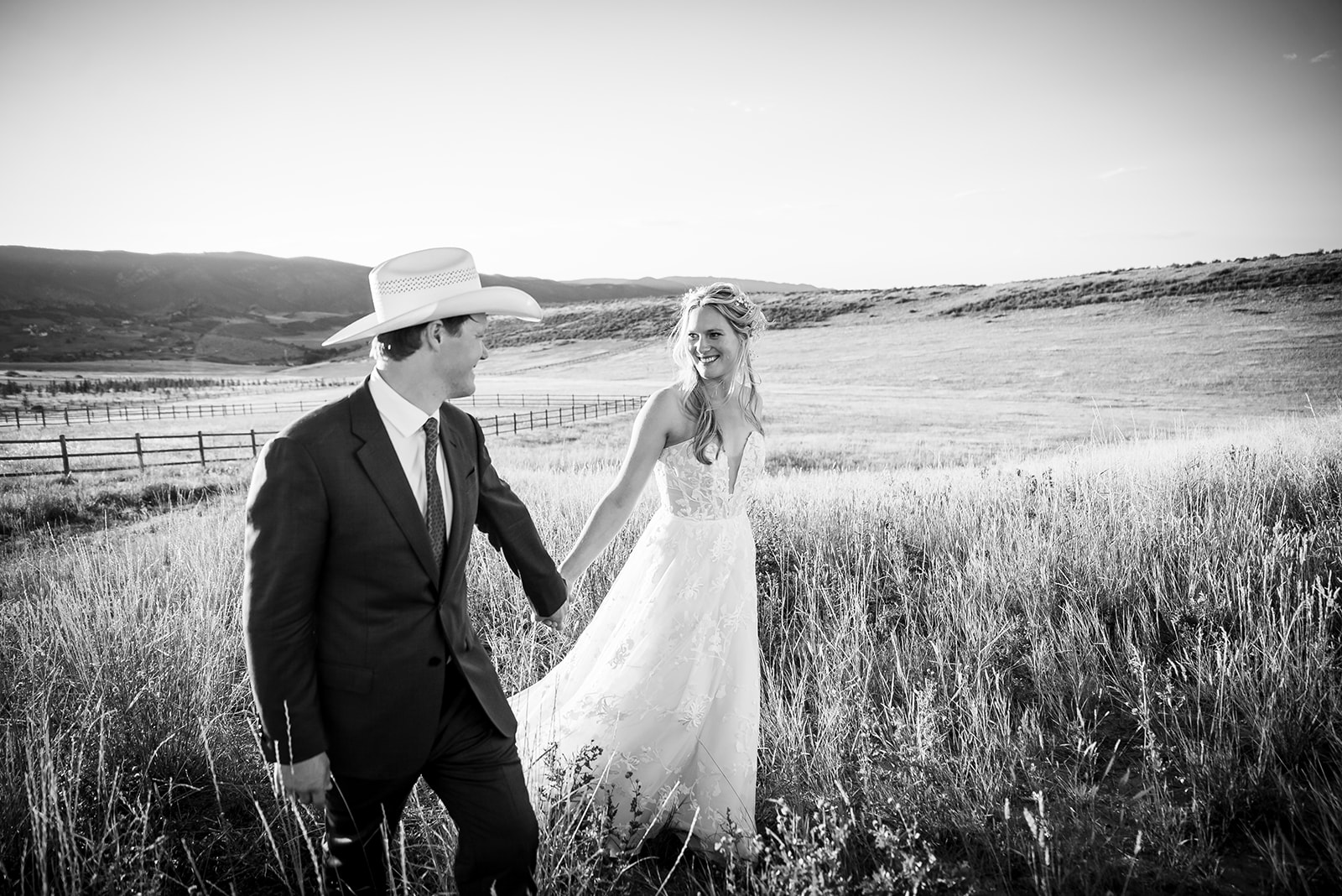 Bride and groom walk through a field hand-in hand looking into each other's eyes.