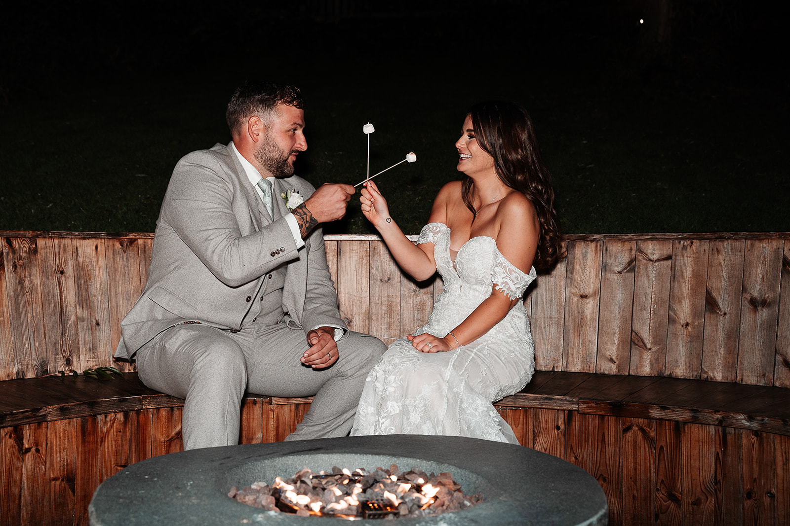 Bride and groom 'cheers' their marshmallows on sticks in front of the firepit at night during their Sandy Cove Hotel wed