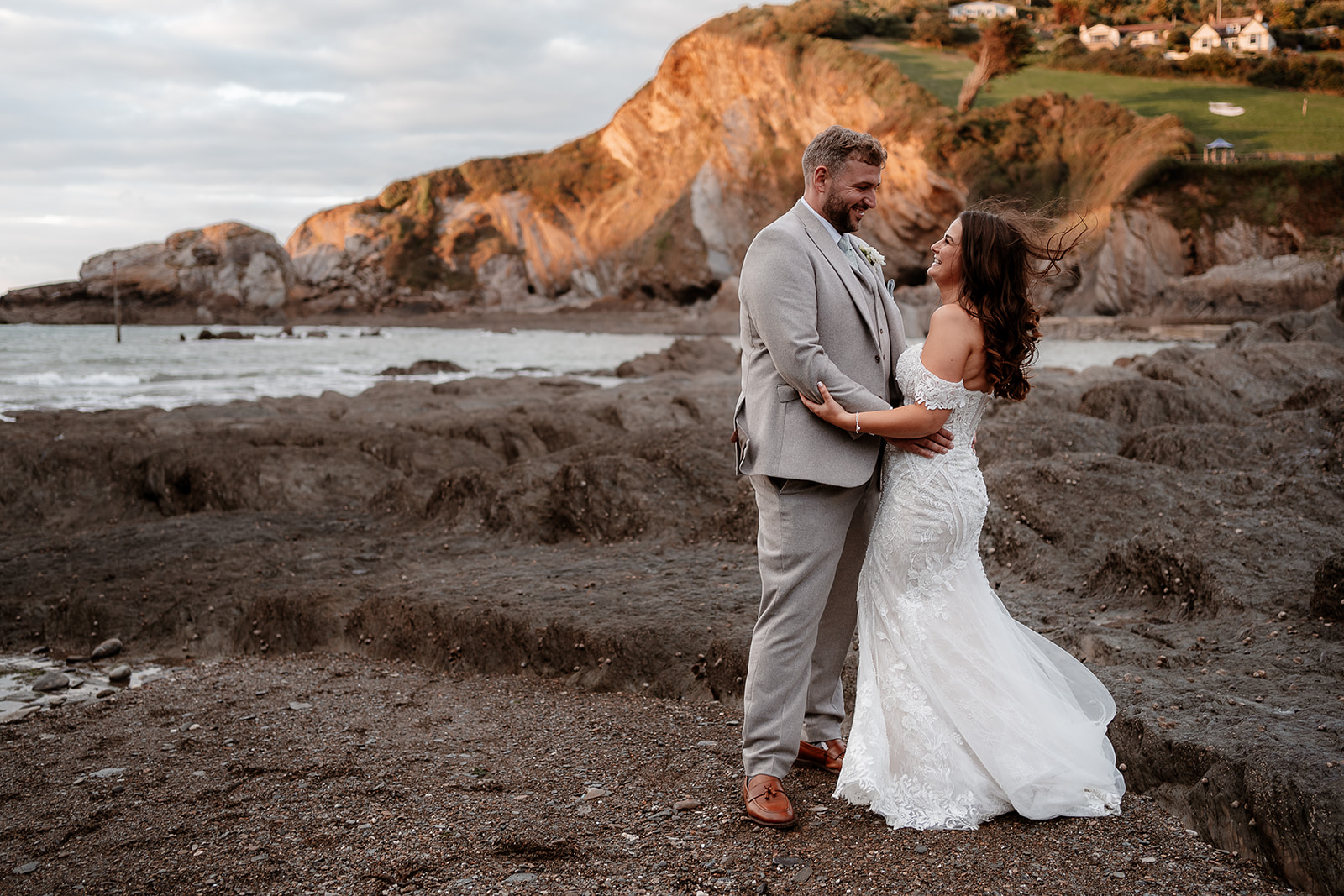 Bride and groom cuddle on the beach with the sun setting on the cliffs behind them at their Sandy Cove Hotel wedding