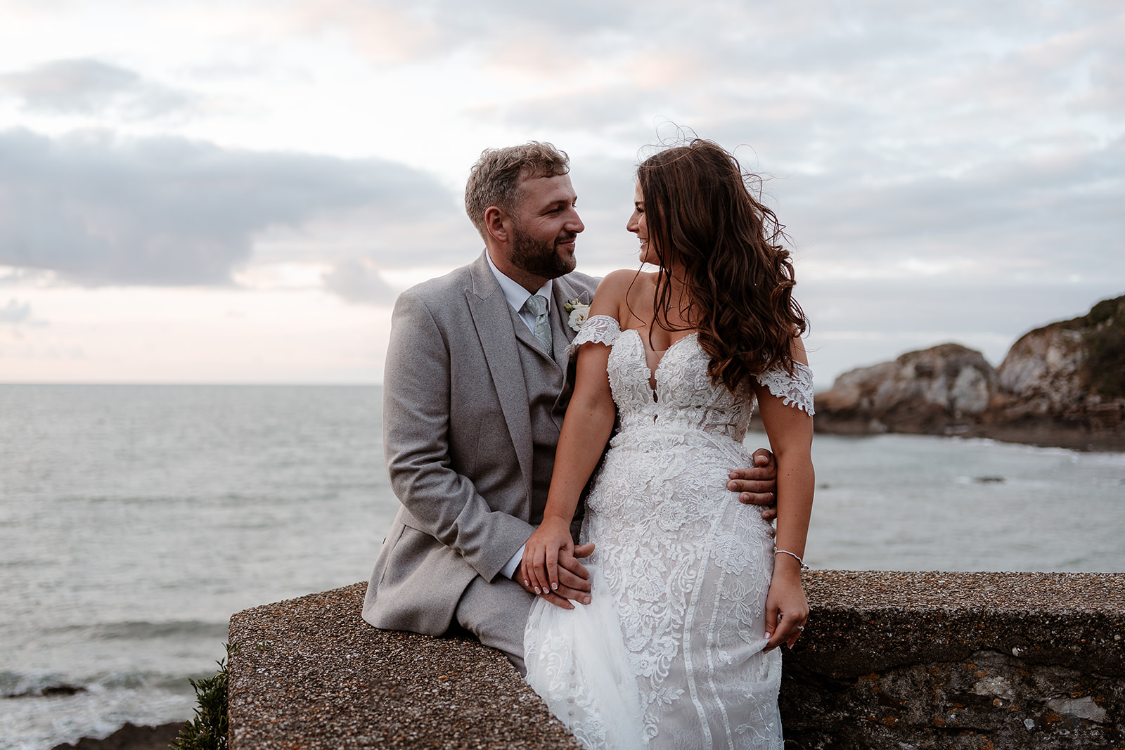Bride and groom cuddle sat on a sea wall at their Sandy Cove Hotel wedding