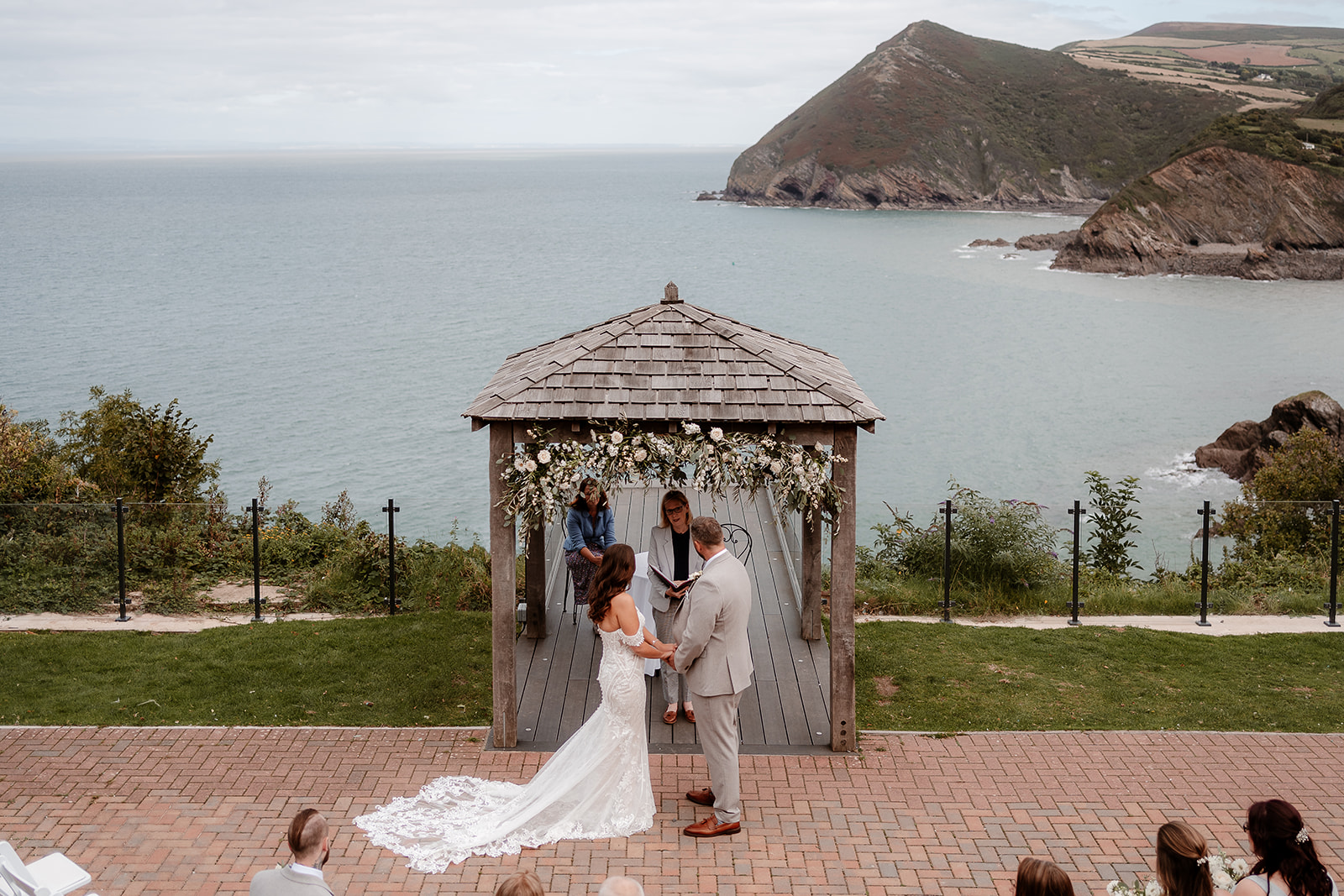 Bride and groom stand under a flower arch in front of the scenic coastal view at their Sandy Cove Hotel wedding