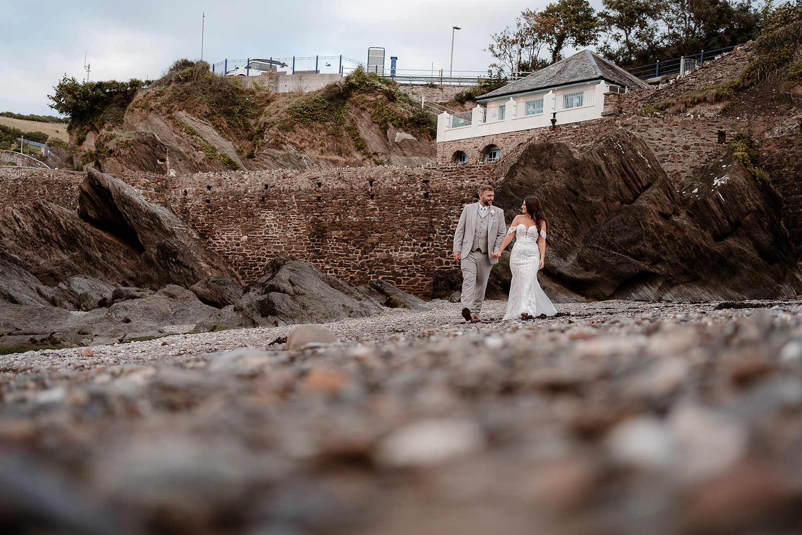 Couple walk hand in hand along a pebble beach at their Sandy Cove Hotel wedding