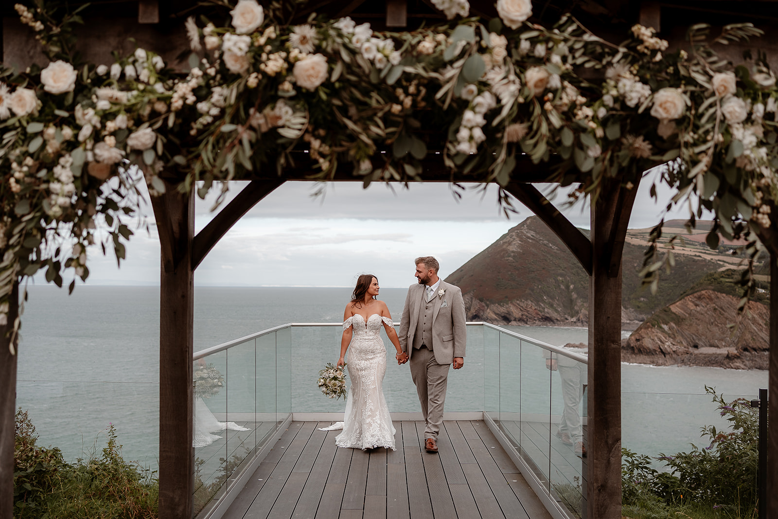 A newlywed couple walk along the platform with the view of the coast behind them at their Sandy Cove Hotel wedding