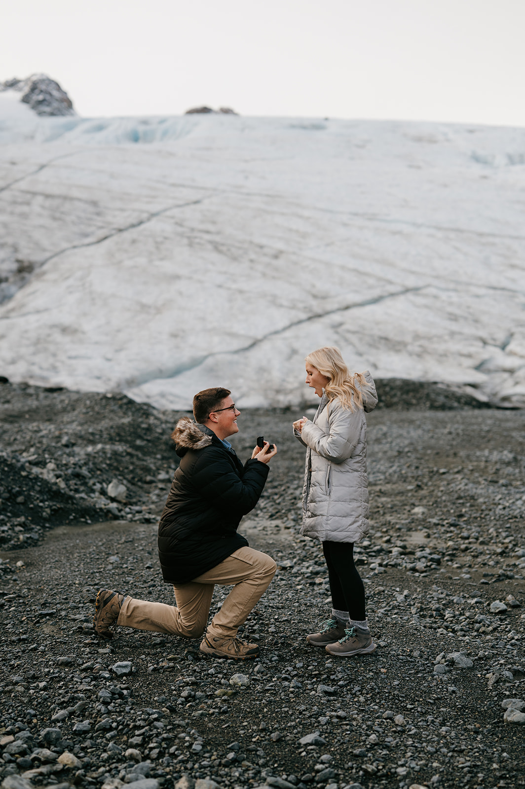 Man gets down on one knee to propose in front of a glacier.